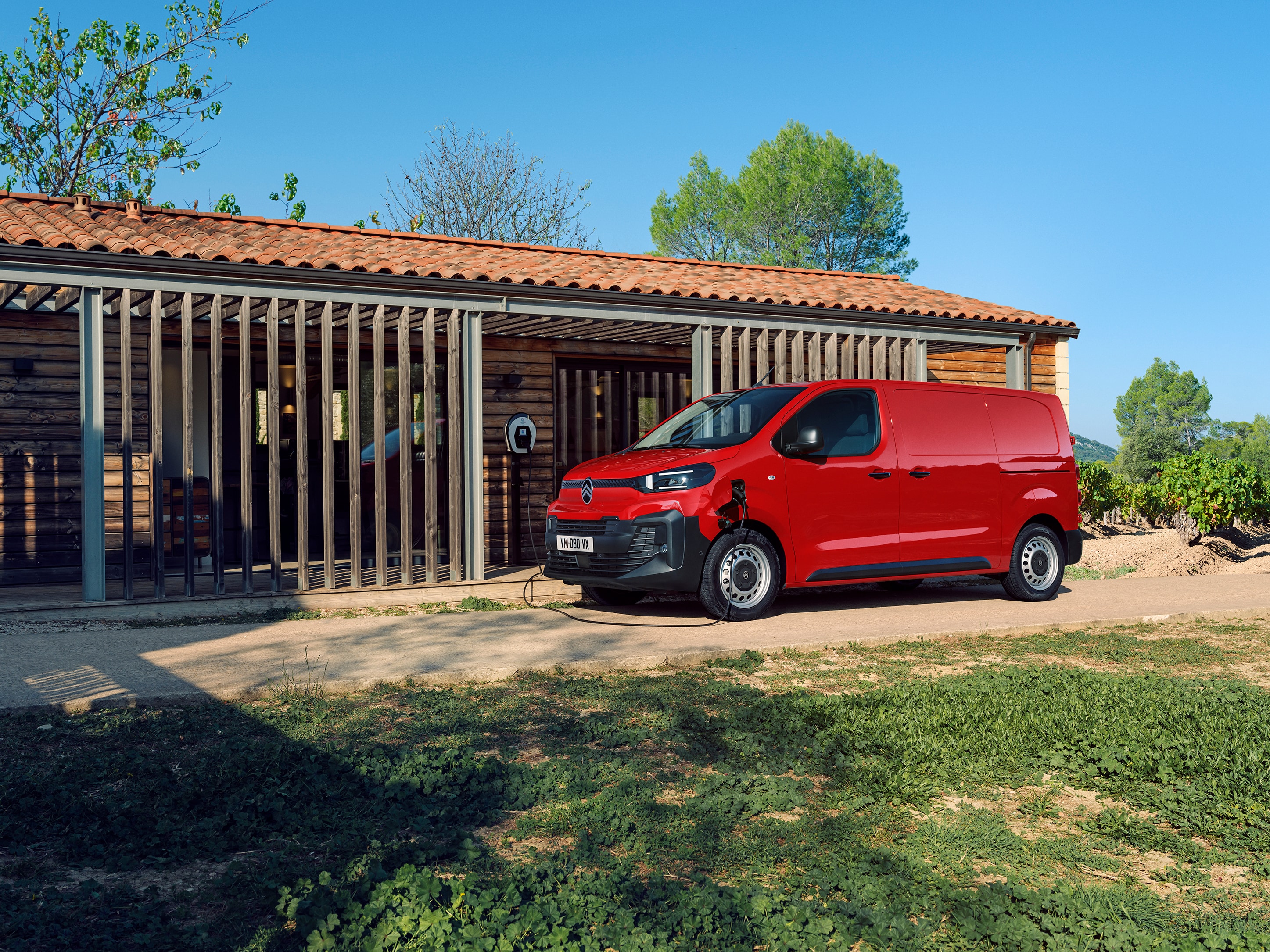 Seitliche Dreiviertel-Außenansicht eines roten Citroën ë-Jumpy beim Laden vor einem Gebäude, zeigt Elektromobilität, Alltagstauglichkeit und professionelles Auftreten.