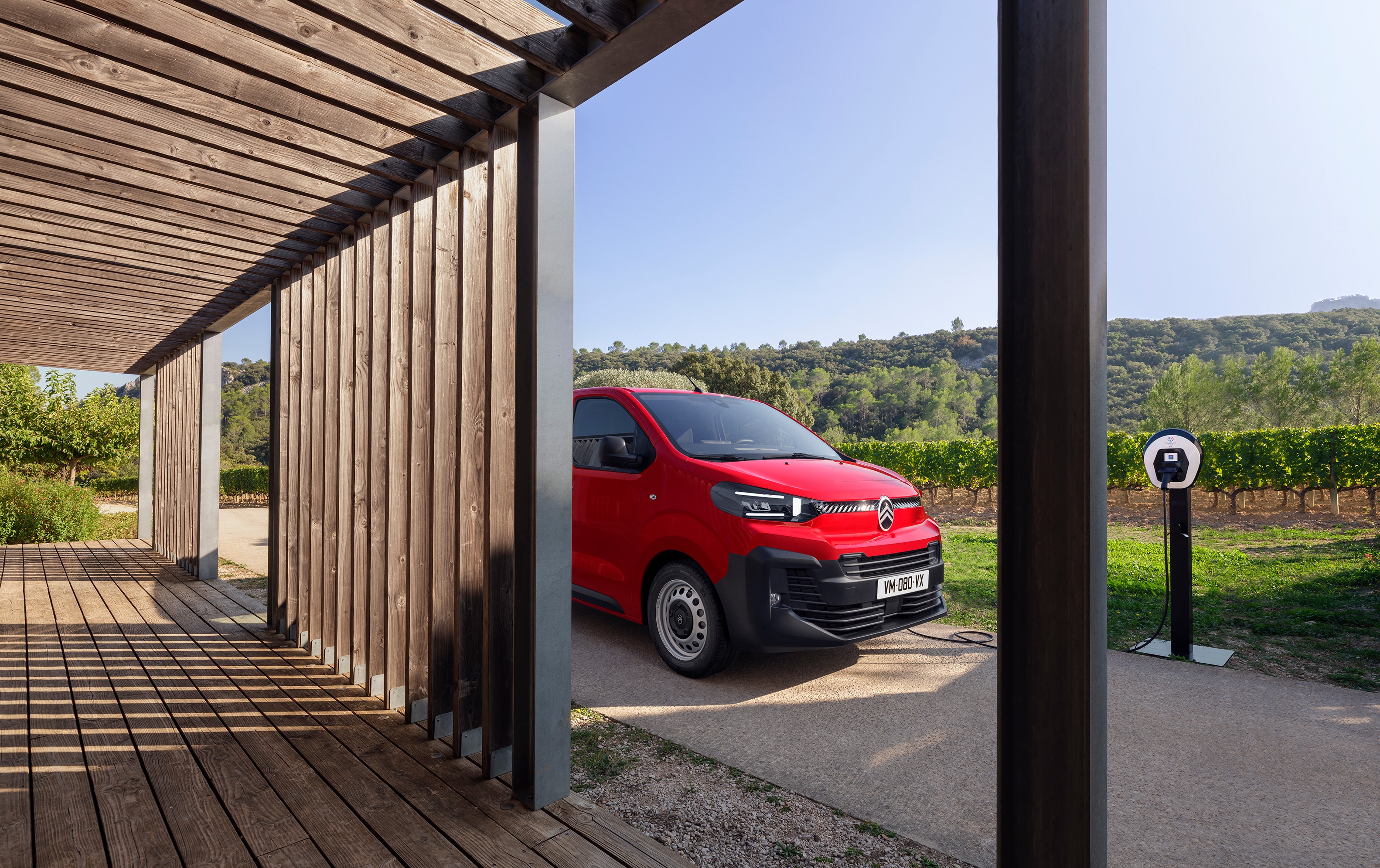Seitliche Dreiviertel-Außenansicht eines roten Citroën ë-Jumpy vor einem Holzgebäude geparkt, zeigt modernes Elektro-Design, Alltagstauglichkeit und professionelles Auftreten.