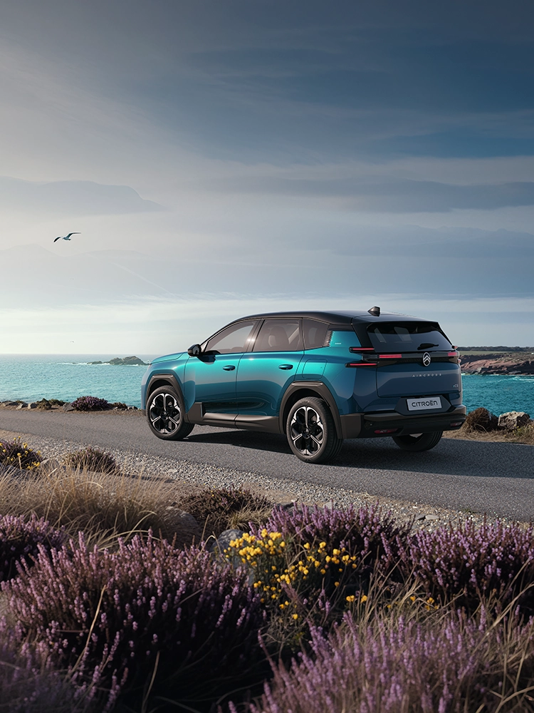 Dreiviertel-Heckansicht des Citroën ë-C5 Aircross Emeraude; zeigt die markanten Light-Wings-LED-Heckleuchten des C5 Aircross auf einer Küstenfahrt mit dem Familien-SUV